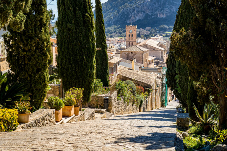 Treppe der Touristenattraktion Calvari Stairway in der Altstadt von Pollensa mit Blick über die Dächer auf den Turm der Kirche Santa Maria dels Àngels und den kleinen Berg Puig de Maria an einem sonnigen Frühlingstag.